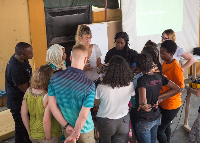A woman in a white t-shirt is surrounded by 10 others looking on as she opens the medical kit.