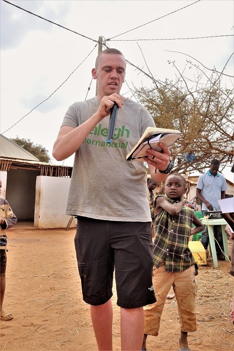 GLIDE employee Joe delivering a speech to community in Matanga on action day with young child in background