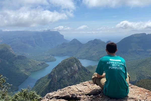 A volunteer sitting on a rugged cliff, gazing out at mountains.